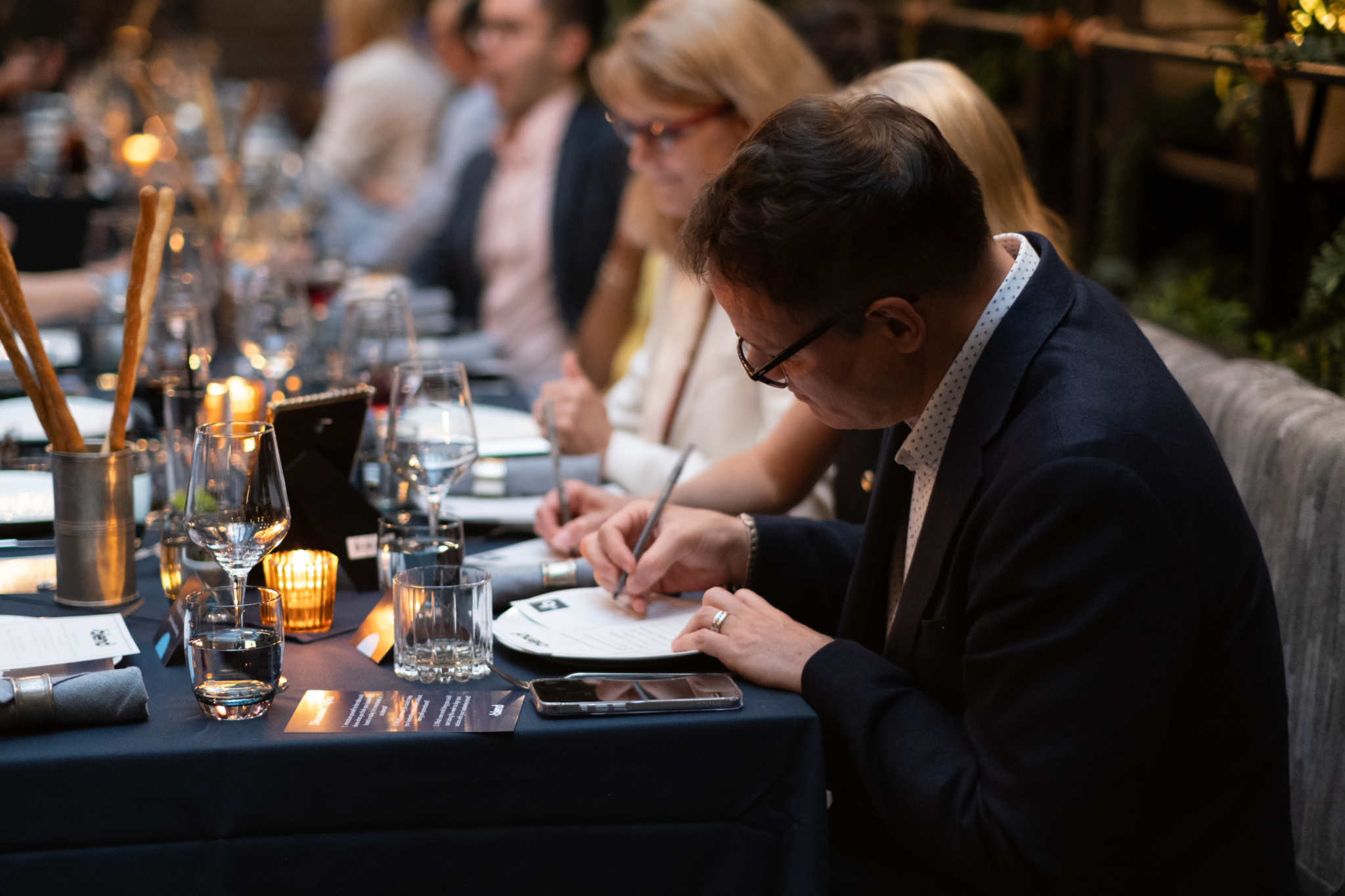 A group of seated C-suite professionals fill in Blend branded forms at a Blend community networking event. They are sat at a long dining table.