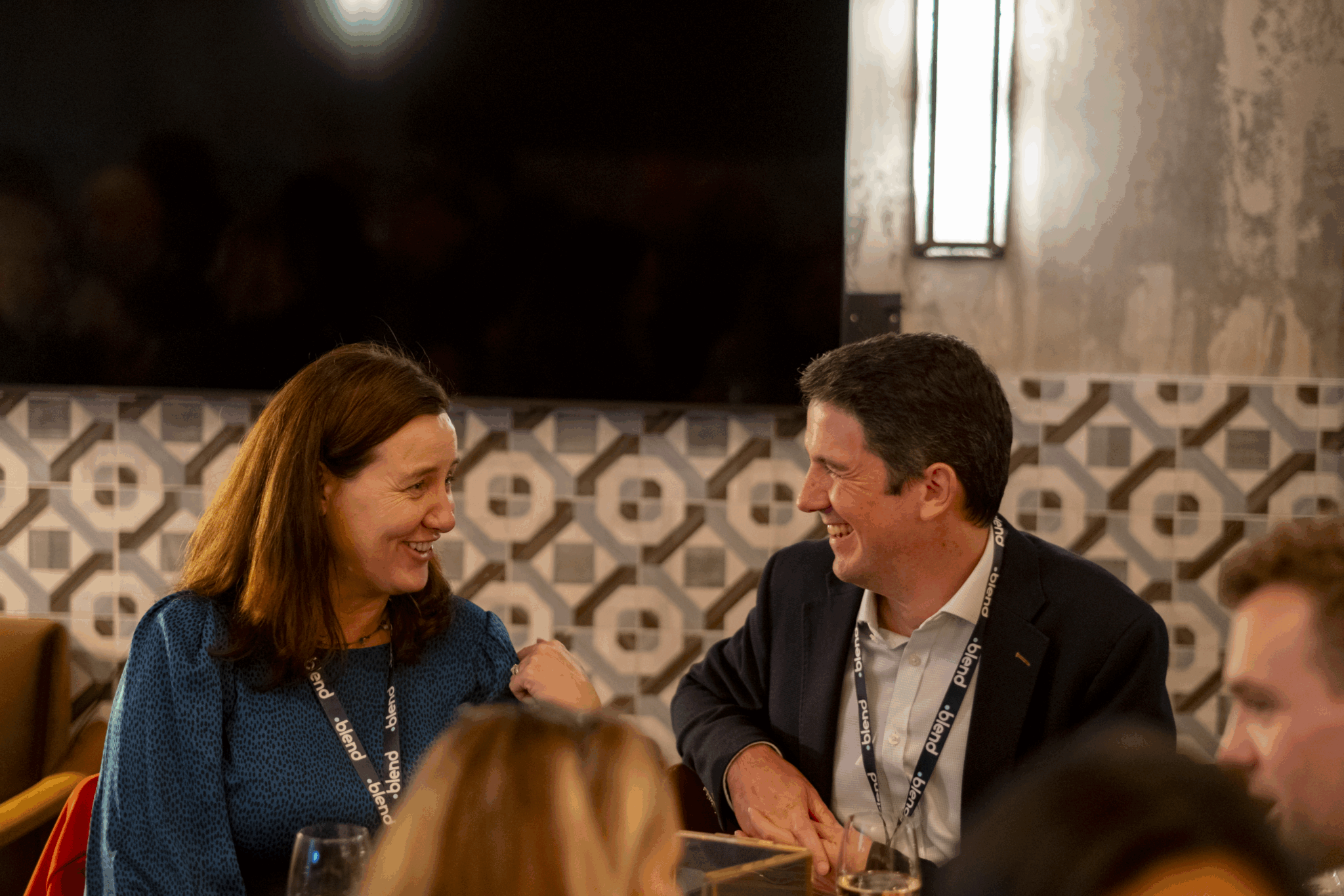 Two C-suite professionals talk at a Blend community networking event, wearing Blend branded lanyards. They are seated.
