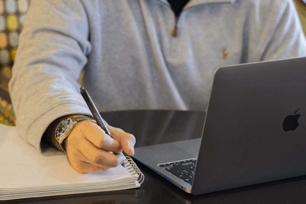 A person takes notes with a notepad and pen whilst working at a laptop