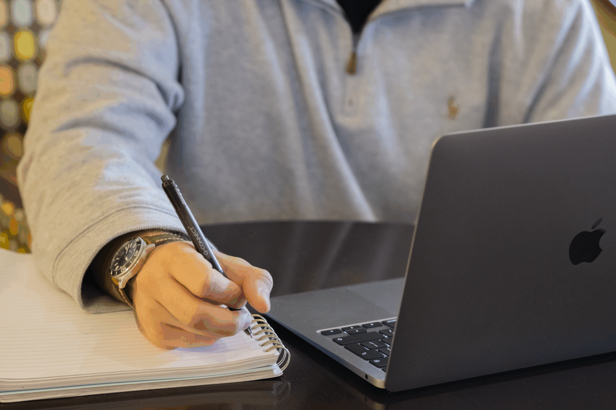 A person takes notes with a notepad and pen whilst working at a laptop